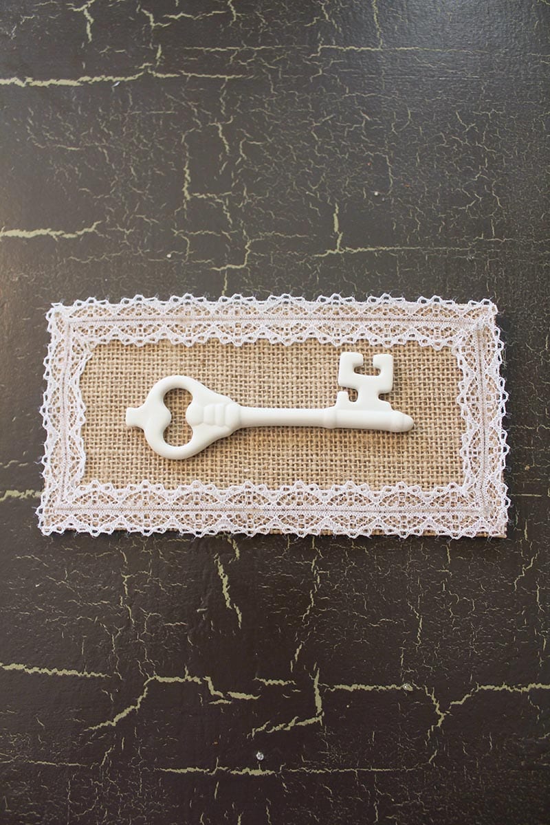 Key Artwork | A white porcelain key, burlap backing and lace all come together in this vintage-inspired mounted key artwork. A super easy DIY for a super cute decor item!