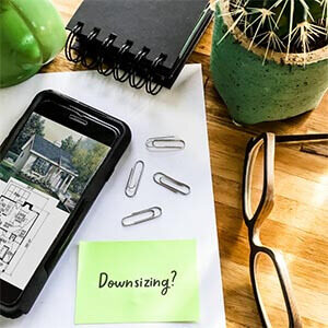 Office supplies such as paperclips and a pen sitting on a wood table with a plant, a pair of glasses and a tape measurer. A green note with the word "downsizing?" sits next to a phone with a floor plan on the screen. Click to visit post.