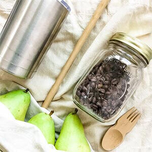 A stainless steel coffee mug, a bamboo straw, a bamboo fork, a glass jar and green pears sitting on a fabric grocery bag. Click to visit post.