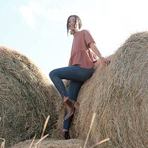 A woman sitting on a hay bale wearing jeans and ankle boots.