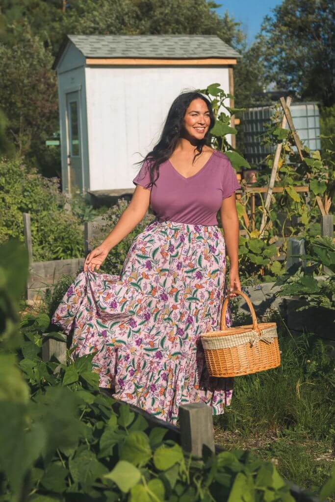 A woman in a garden wearing a purple tee and a flowing floral skirt.